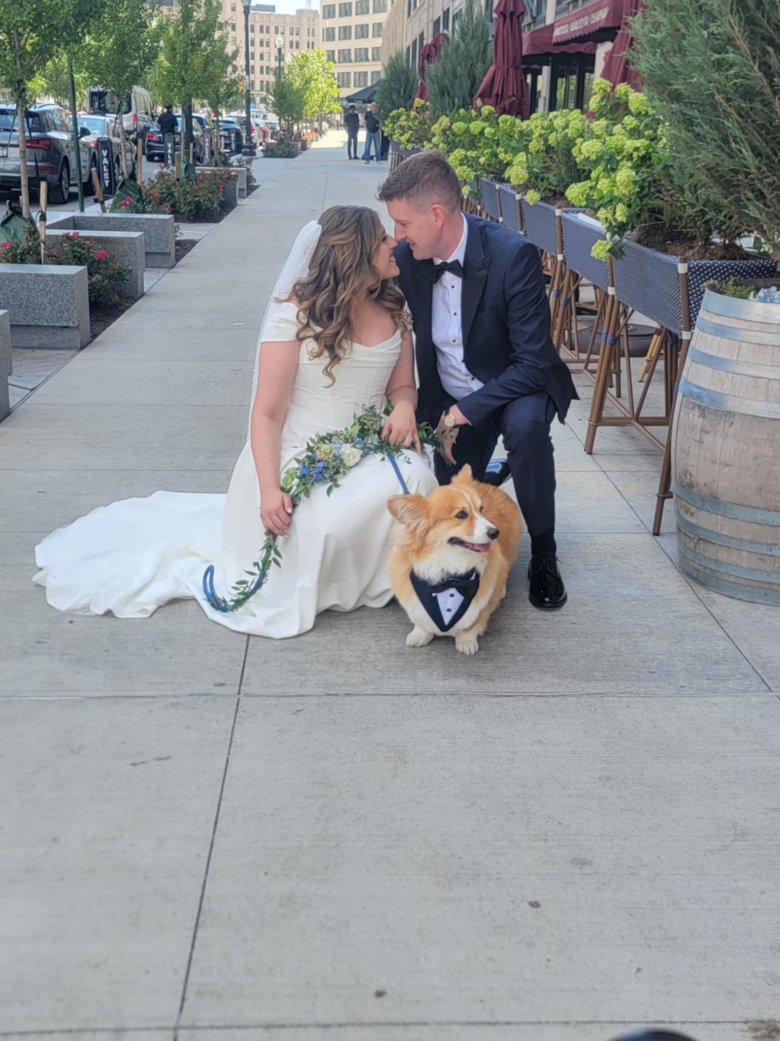 Bride and groom kneel with a dog on a city sidewalk, surrounded by planters and outdoor seating.