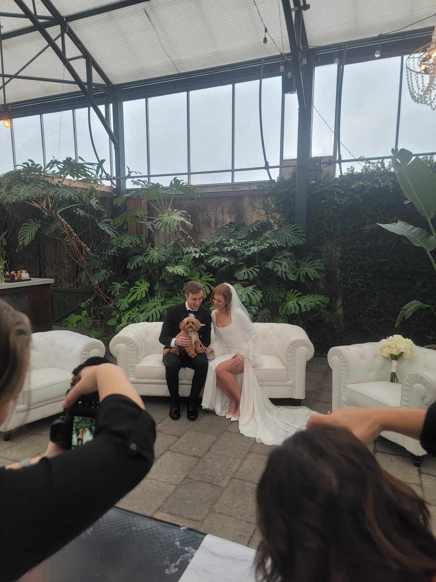 A bride and groom sit on a white sofa with a child, inside a greenhouse with plants and glass ceiling.