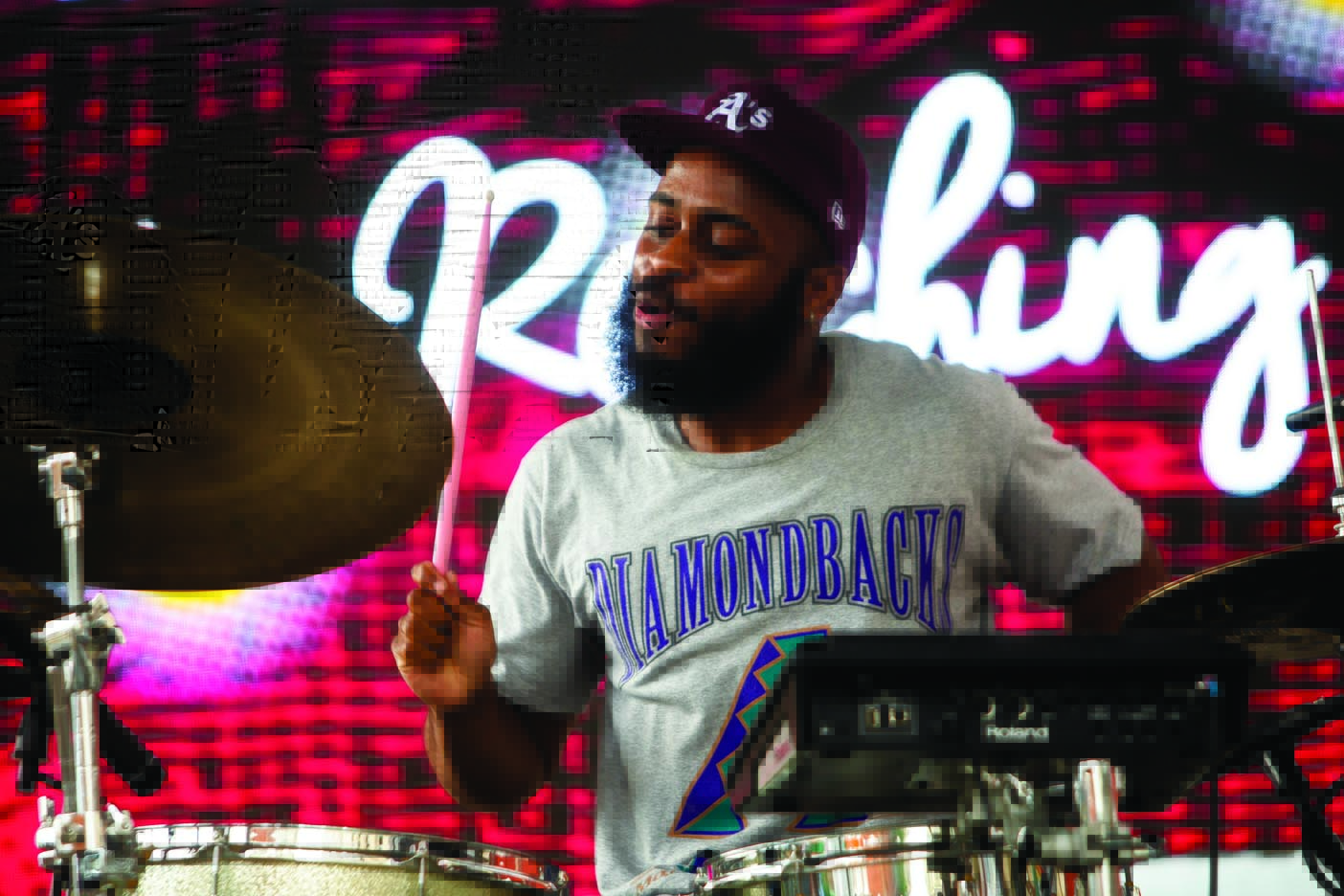 A man playing drums on stage with a red and black background, wearing a gray shirt and a black cap.