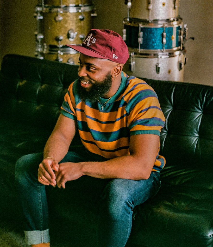 Man with a beard wearing a red cap and striped shirt, sitting on a black couch, smiling, with drums in background.