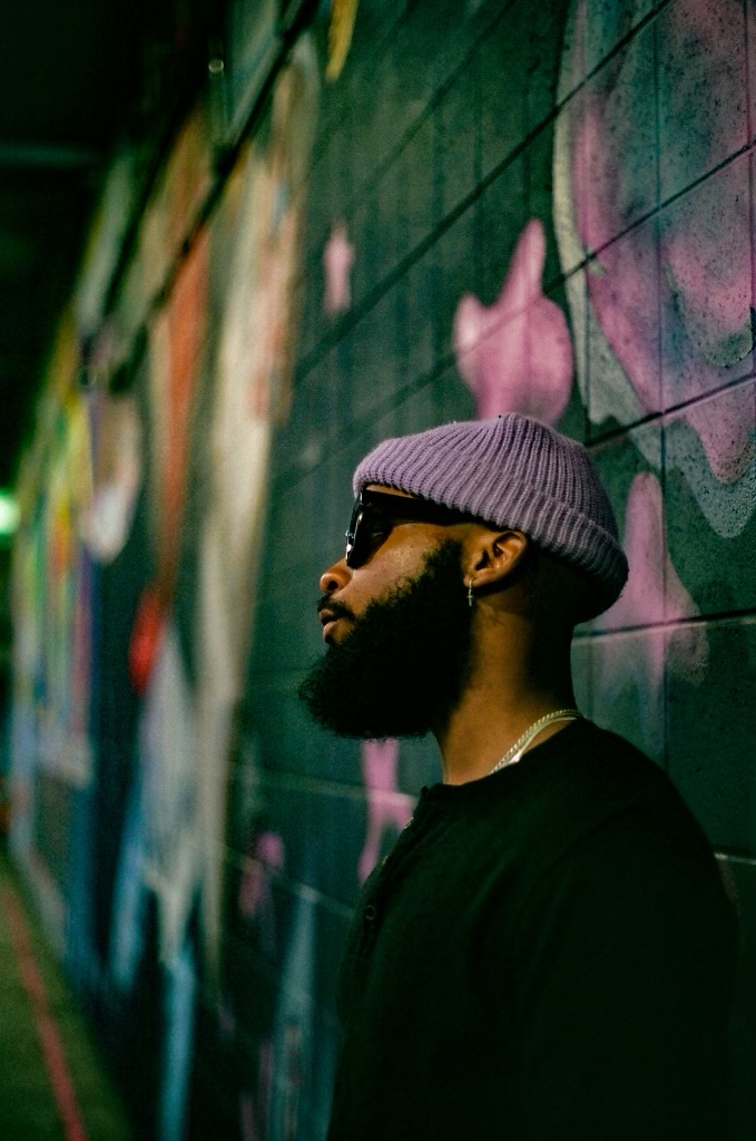 Side profile of a man with a beard wearing sunglasses, a gray beanie, and a black shirt, standing against a colorful graffiti wall.