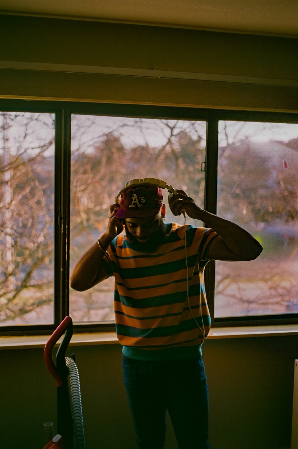 Person adjusting cap in front of large window with trees outside, indoors, wearing striped shirt and jeans.