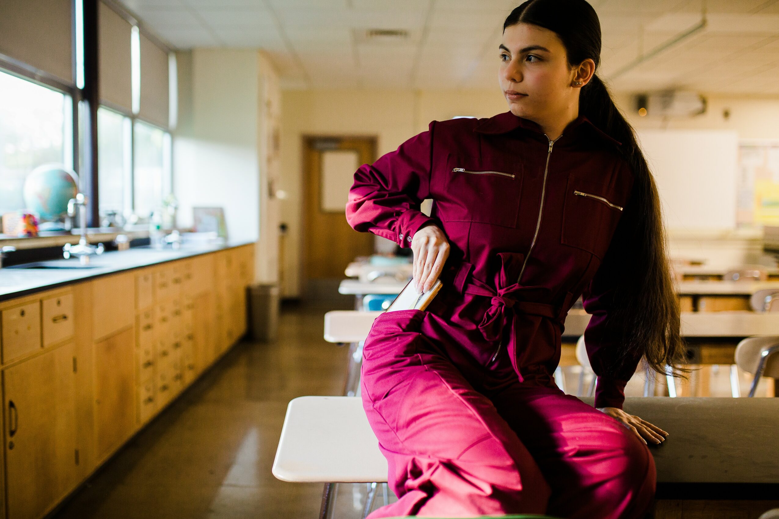 Young woman in a magenta jumpsuit sitting on a table in a classroom or laboratory, with cabinets and windows in the background.