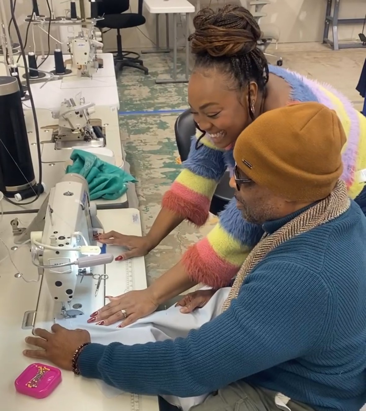 Two women in a laboratory, working with scientific equipment, smiling and engaging with each other, one wearing a colorful sweater and the other a brown hat.