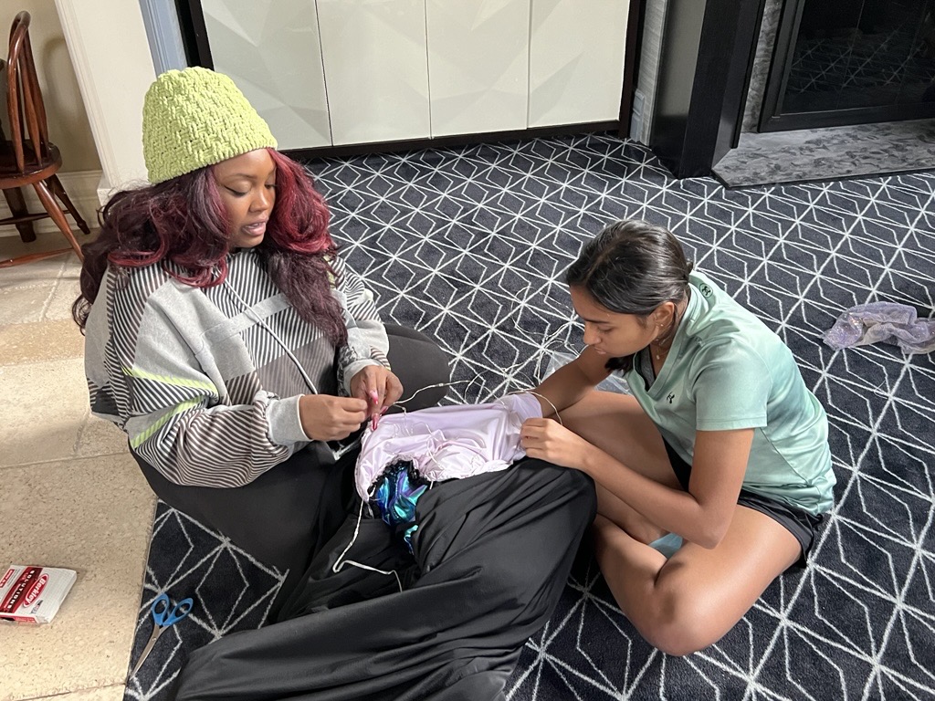 Two women sitting on a patterned floor, looking at a bag with clothes inside, near a white cabinet and a glass door.