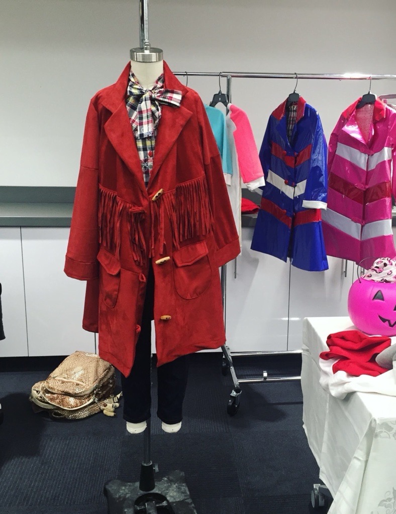 Red coat with fringe, plaid shirt, and dark pants on a mannequin, with colorful children's clothing in the background.