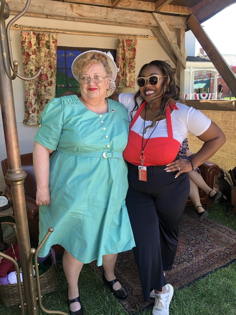Two women standing outdoors, one in a turquoise dress and the other in a red and white top with sunglasses, smiling.