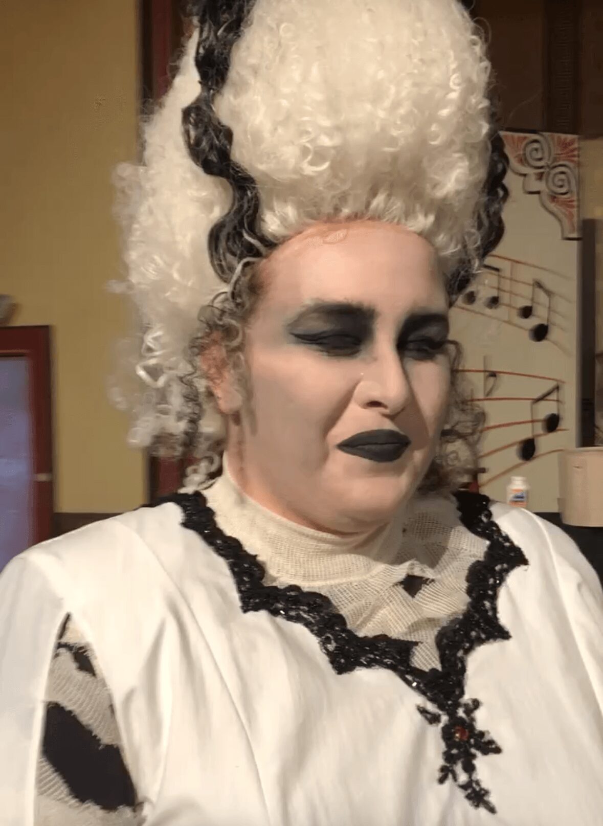 Person with elaborate white wig, dark makeup, and vintage-style clothing, looking downward.