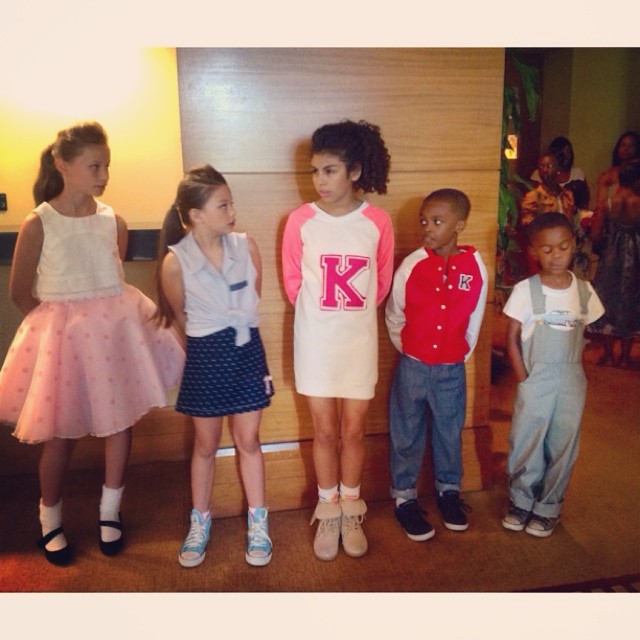 Five children stand in a line indoors, dressed in colorful clothing, with a wooden wall behind them.