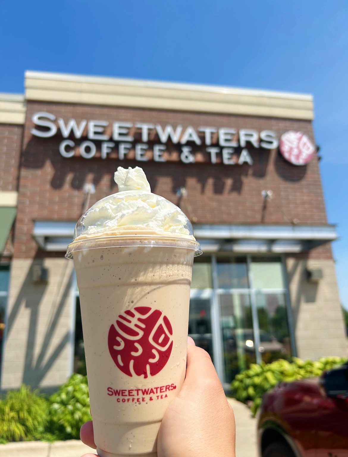 Person holding a blended coffee drink with whipped cream in front of a coffee shop sign, blue sky above.