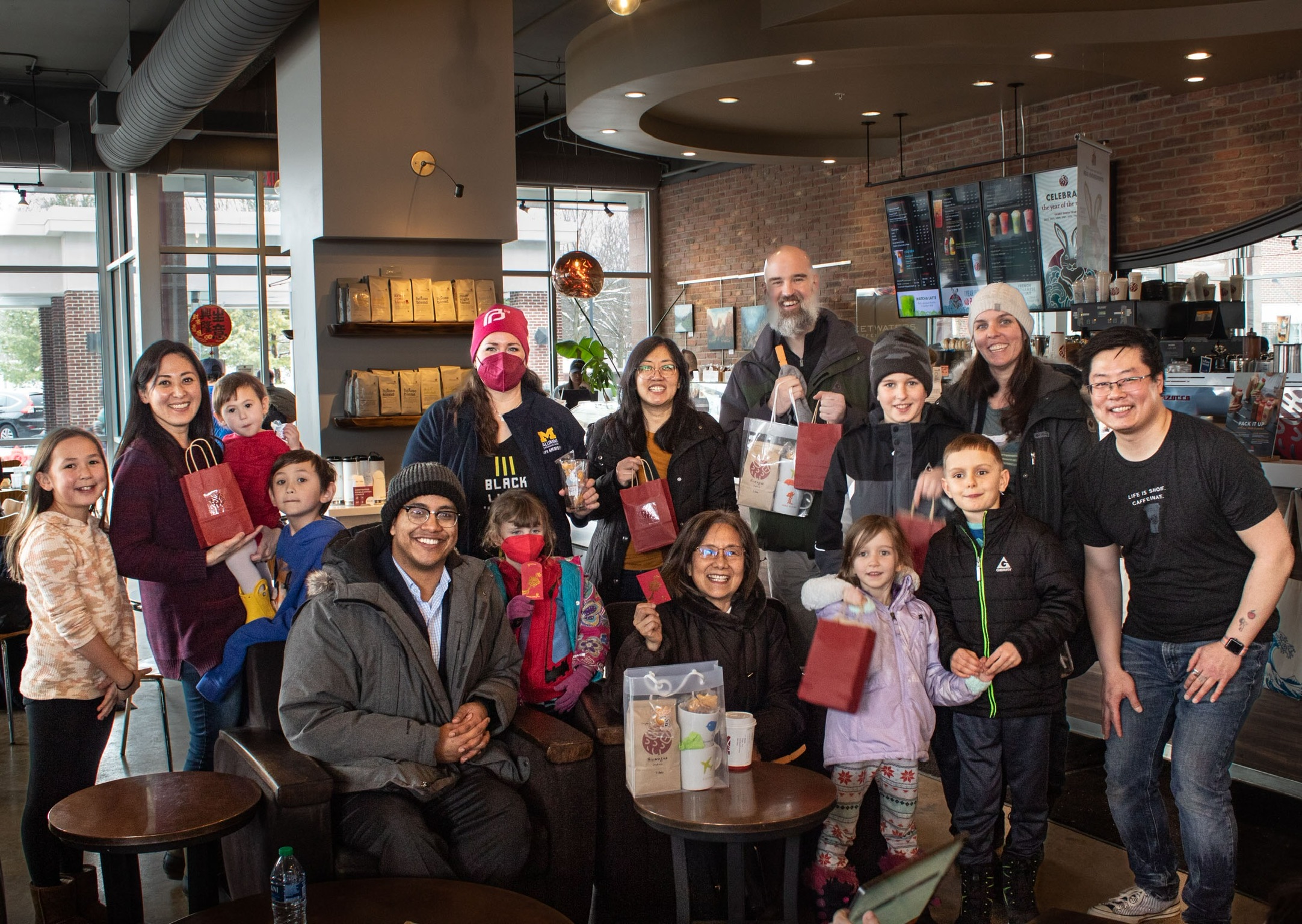 Group of people, including children, gathered in a cafe, some holding gift bags, with a bar and TV in background.