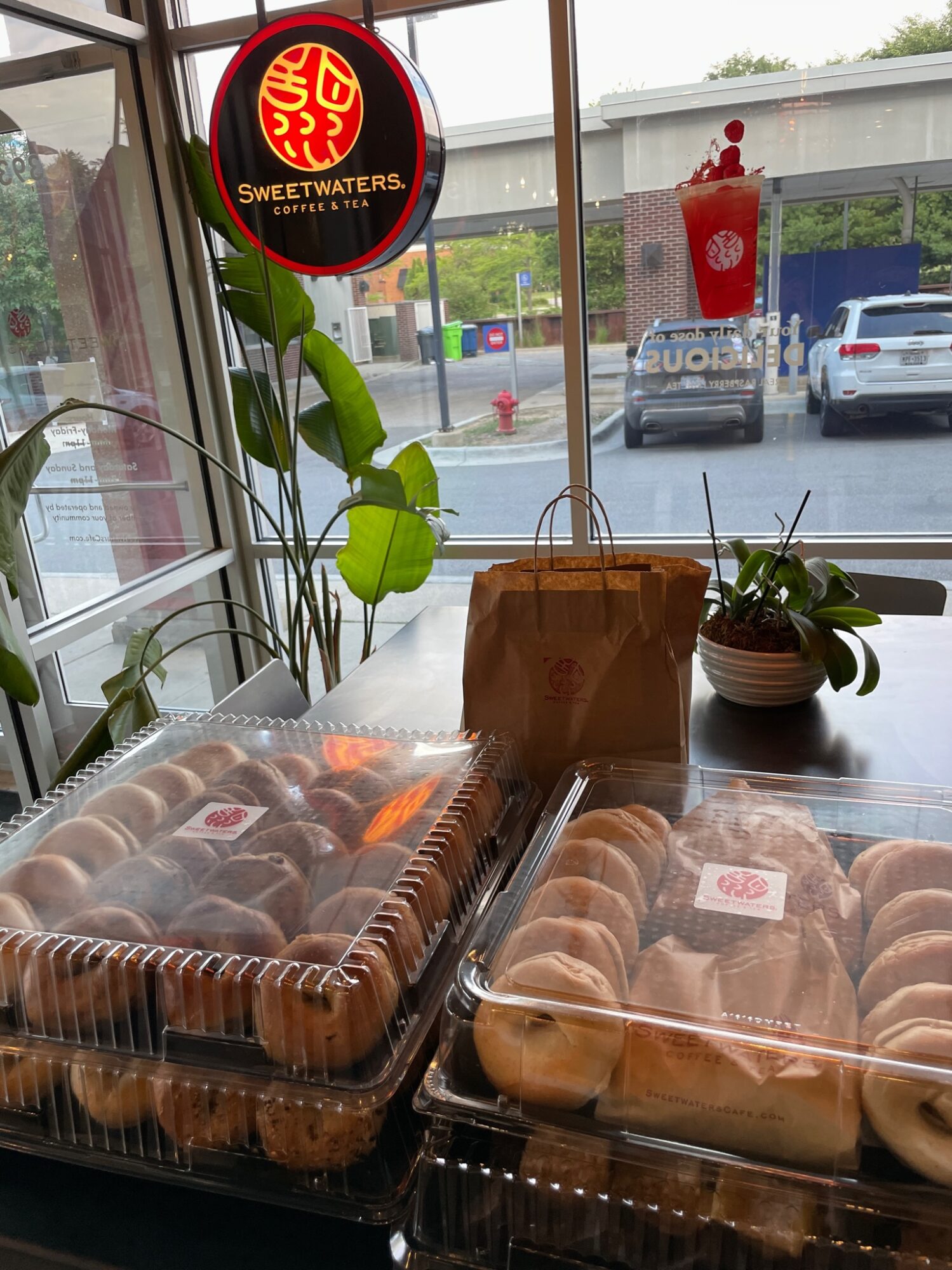 Display of baked goods in plastic containers on a table near a window, with a plant and a Sweetwaters Coffee & Tea sign outside.