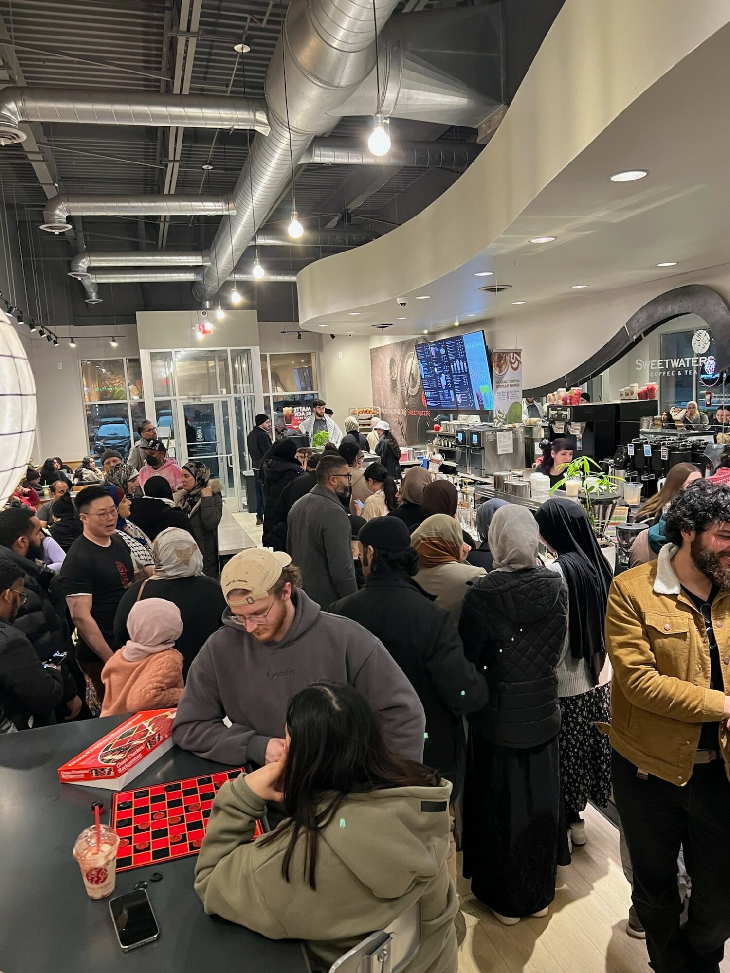 Crowd of people inside a modern shopping mall with a curved ceiling and digital screens, some waiting in line at a counter.