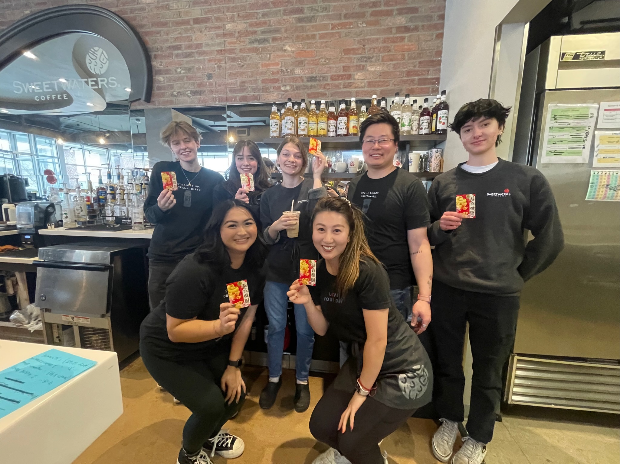 Group of seven people, five women and two men, standing inside a cafe, holding drinks, smiling, with a brick wall and shelves in the background.