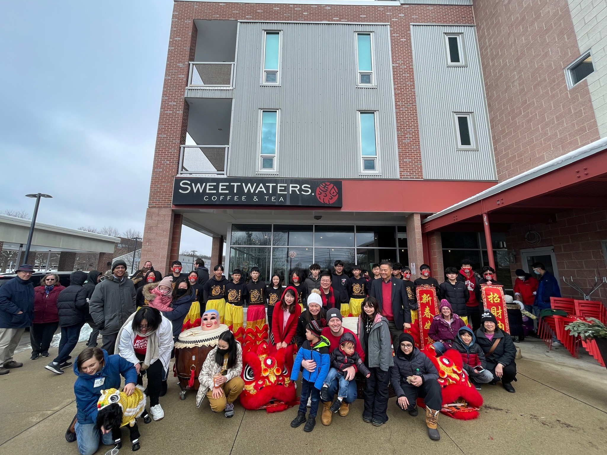 Group of people, children, and adults gathered outside a building with a sign reading 'Sweetwaters Coffee' under a cloudy sky.