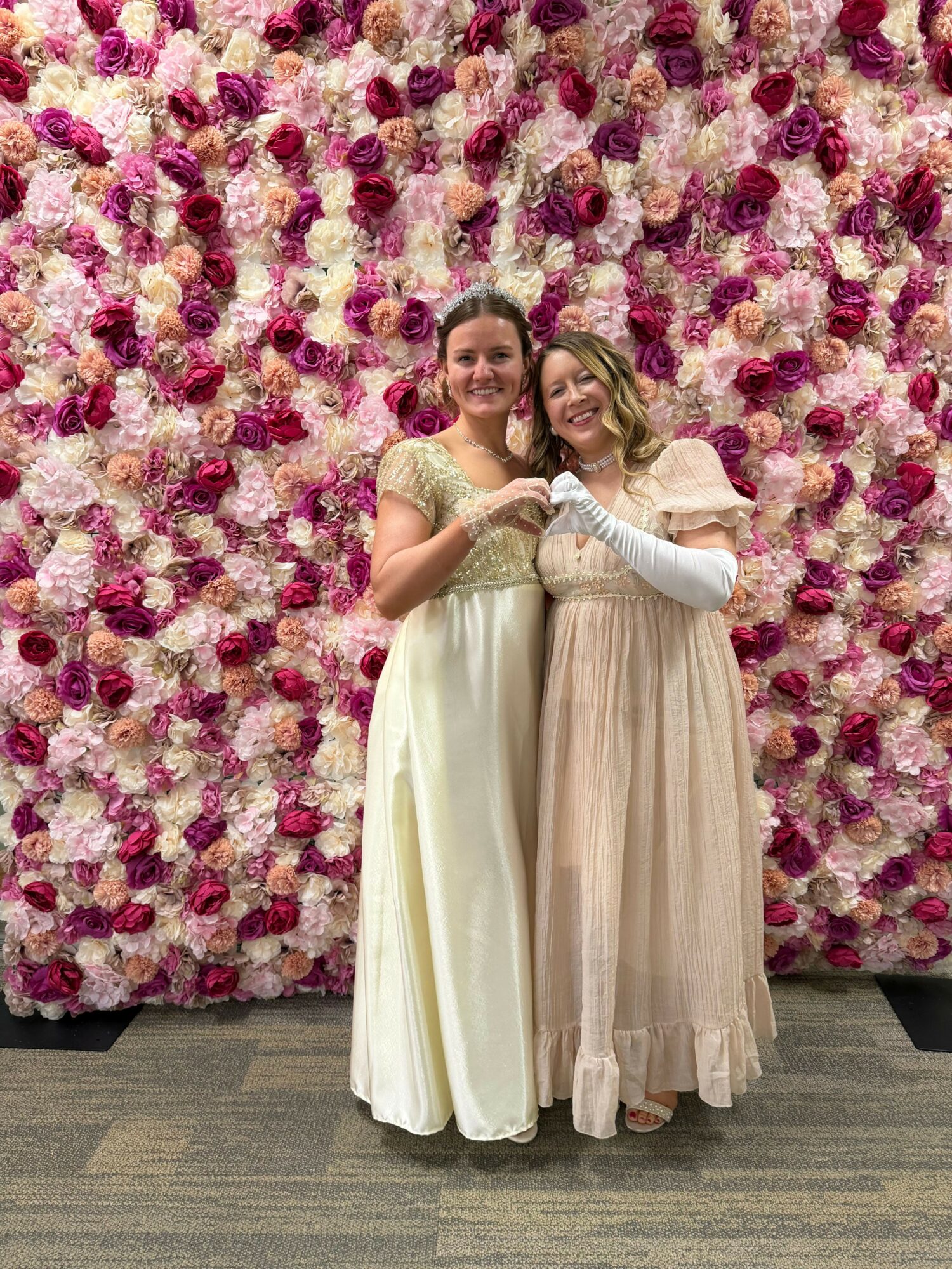 Two women in formal dresses standing in front of a floral backdrop, holding glasses, smiling, at an event.