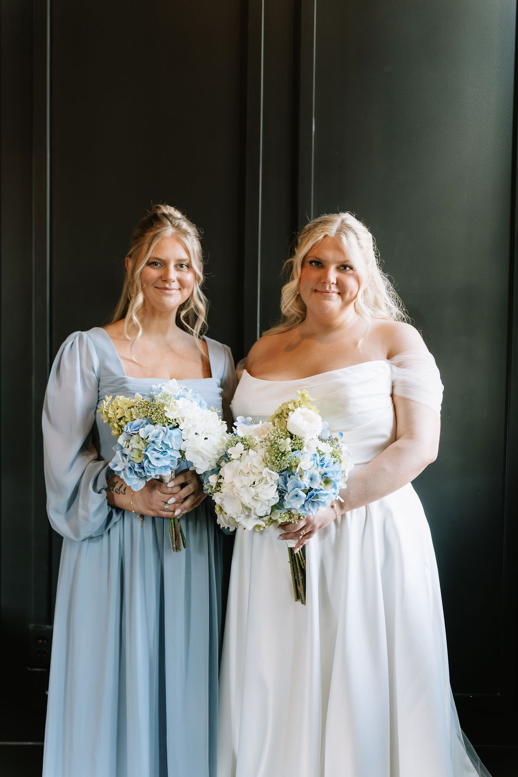 Two women in wedding dresses holding bouquets, standing side by side against a dark background.
