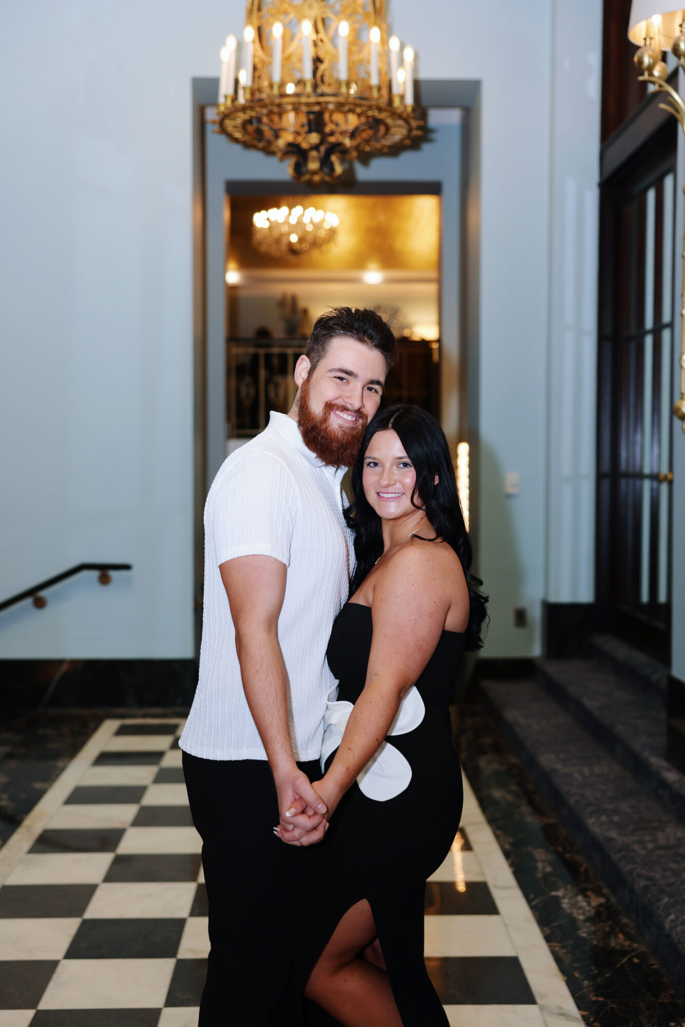A smiling couple holding hands in a grand hallway with chandeliers and checkered floor.