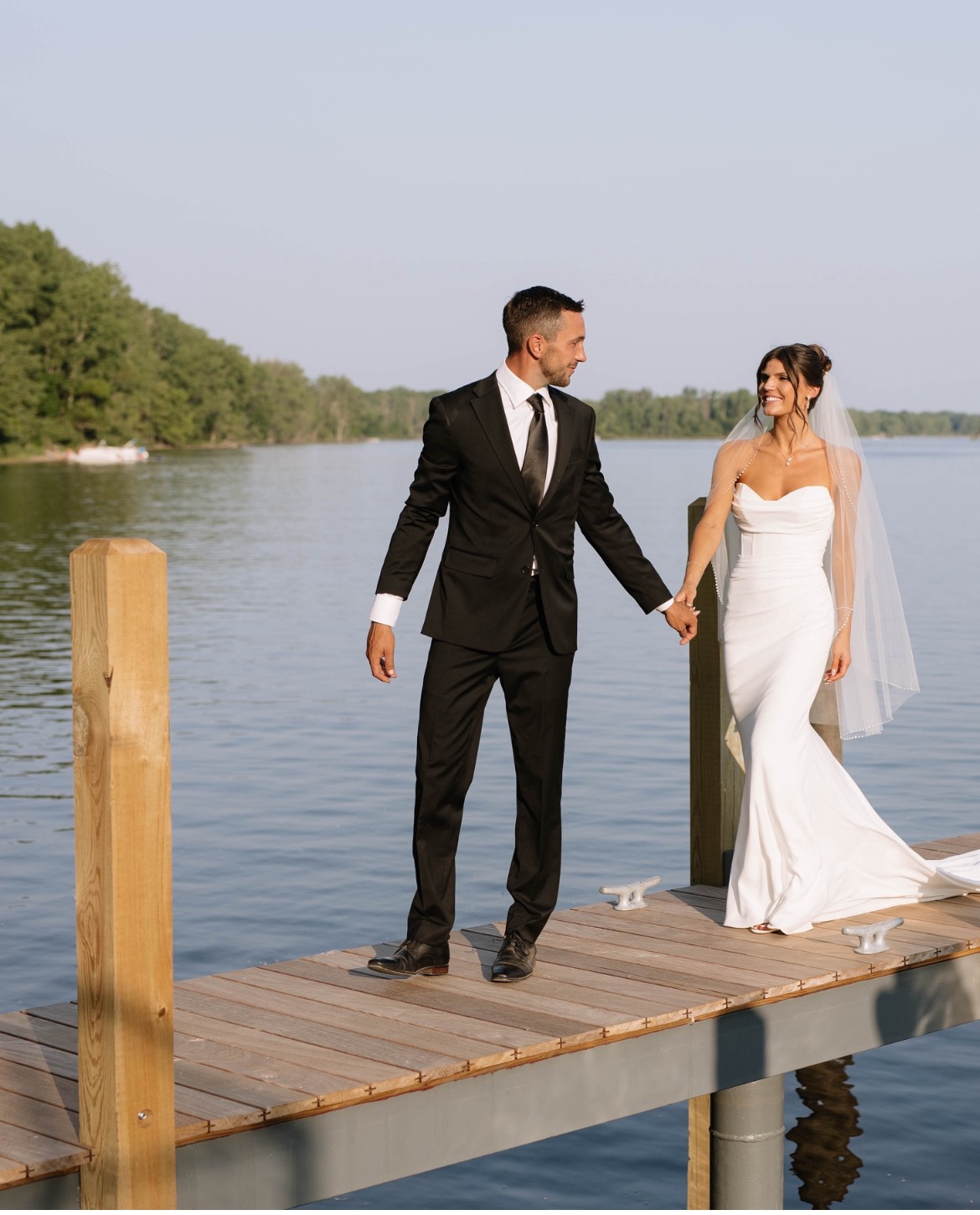 A bride and groom holding hands on a wooden dock by the water, with trees in the background, during daytime.