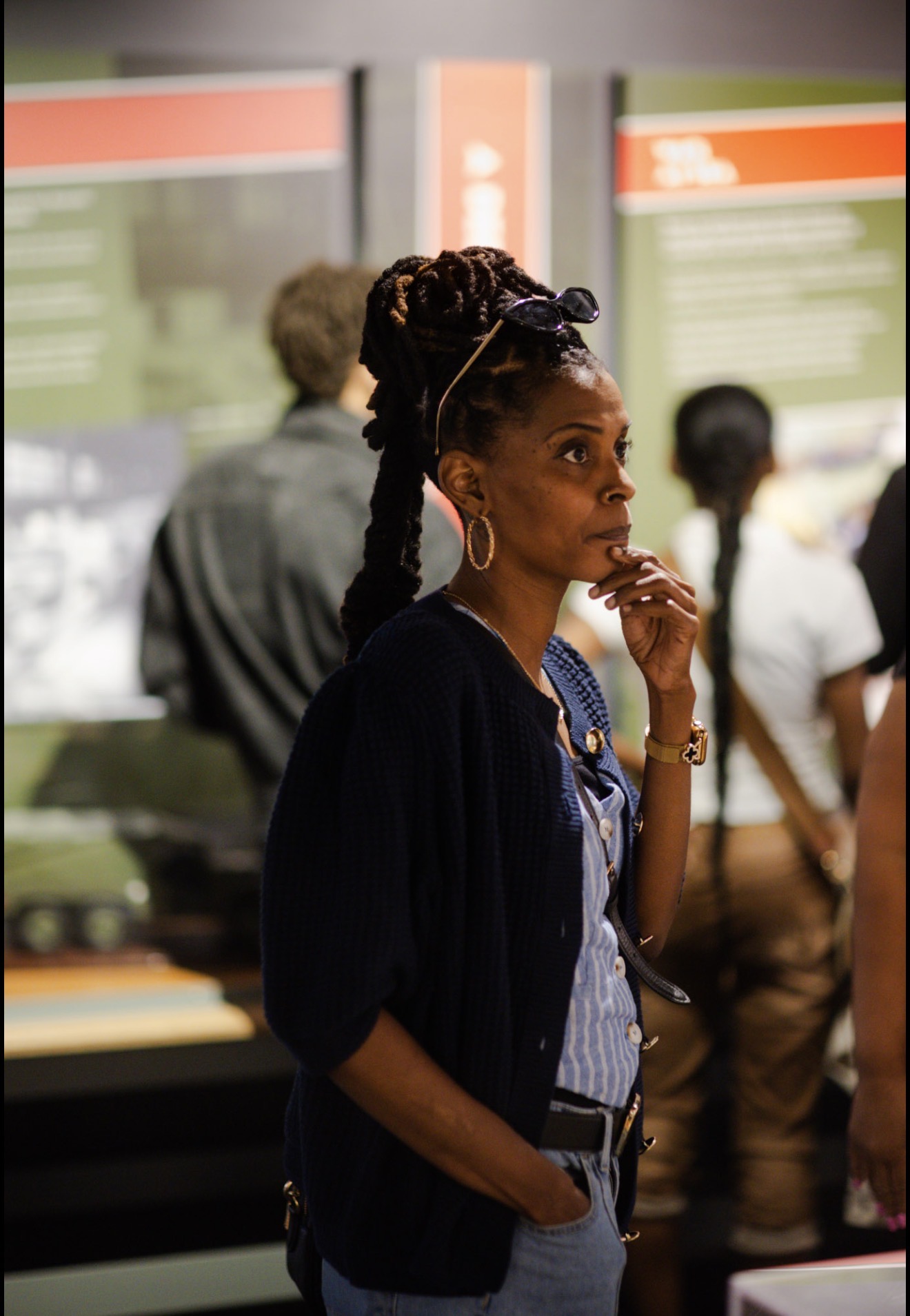 Woman with braided hair and hoop earrings stands in a public space, with people and informational signs in the background.