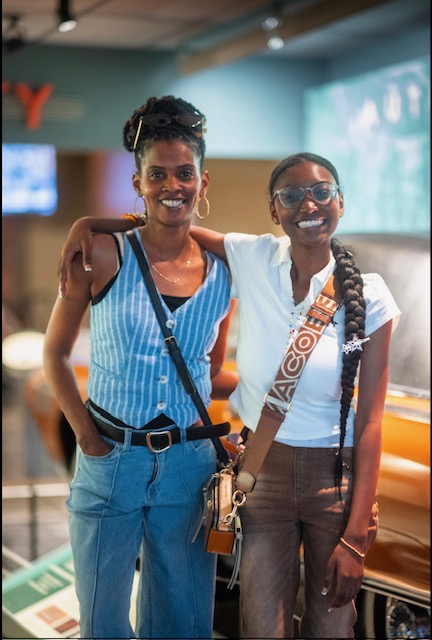 Two women smiling, one with arm around the other, in an indoor setting with a blurred background.