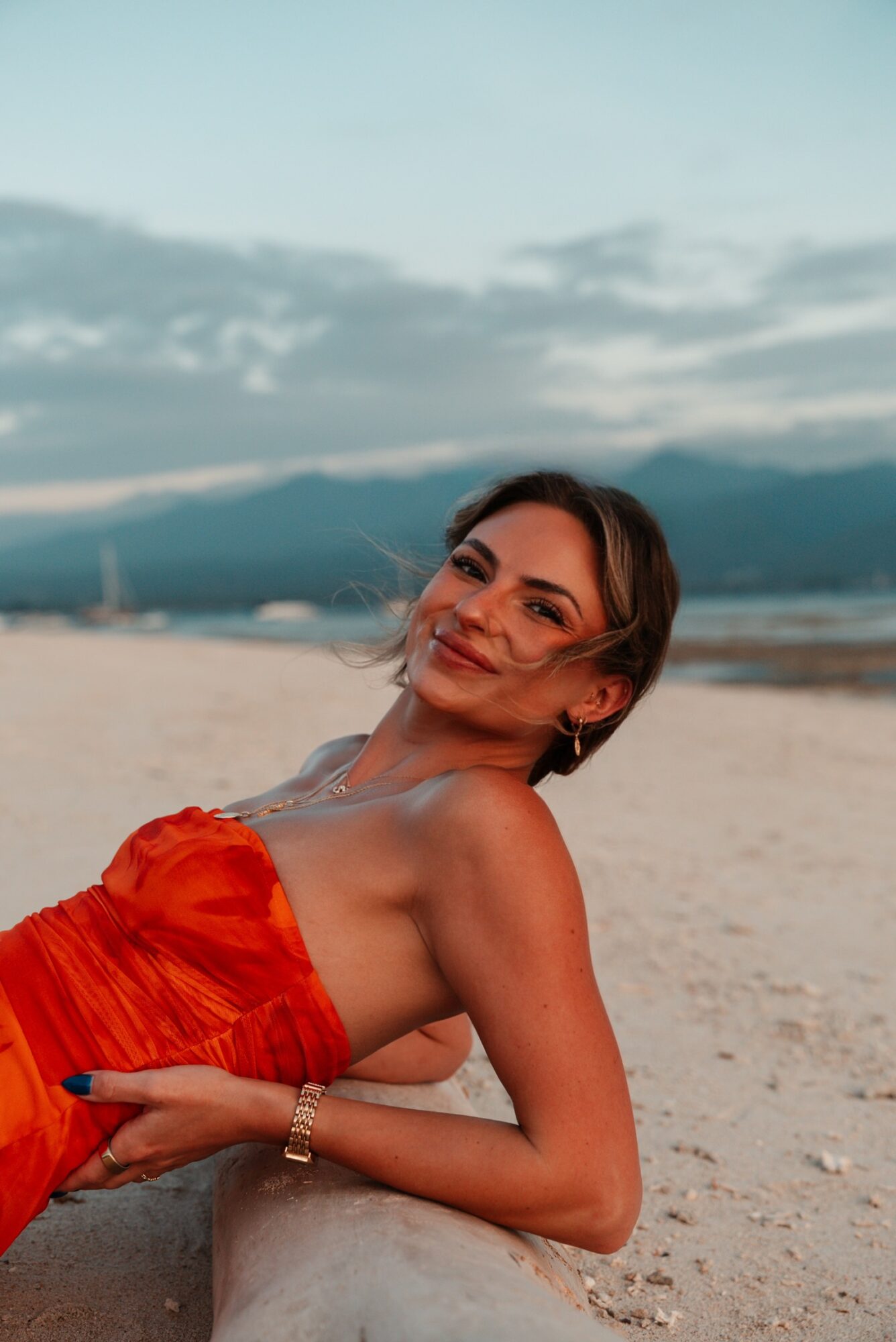 Woman lying on sand beach, smiling, wearing a red strapless dress, with mountains and sky in background.