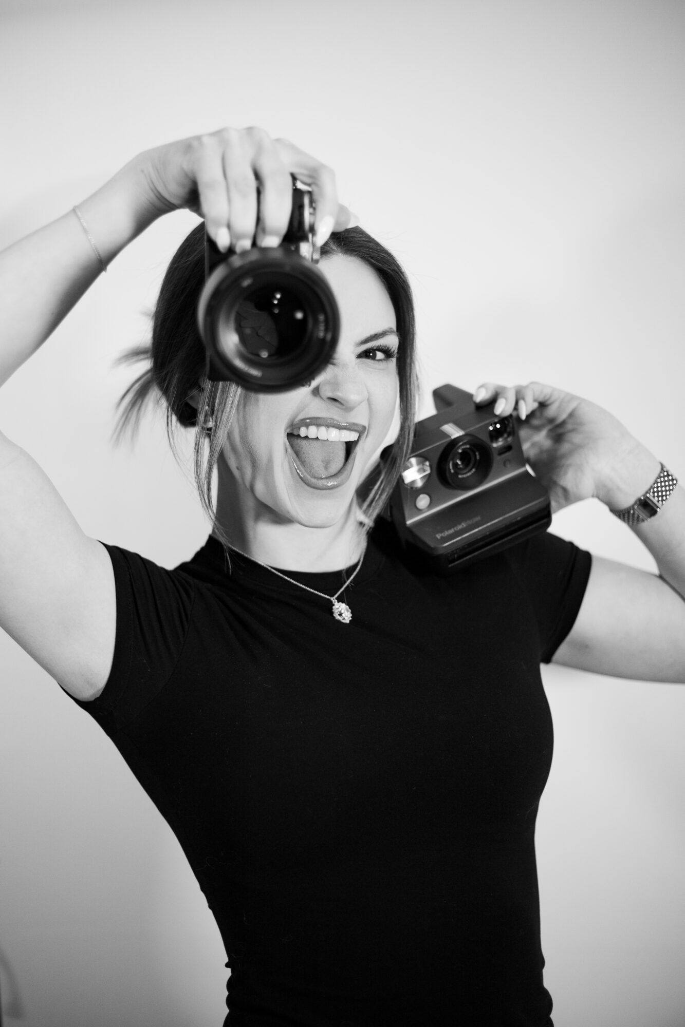 Woman holding two cameras, smiling, with one eye open, wearing a black shirt and necklace, against a plain background.