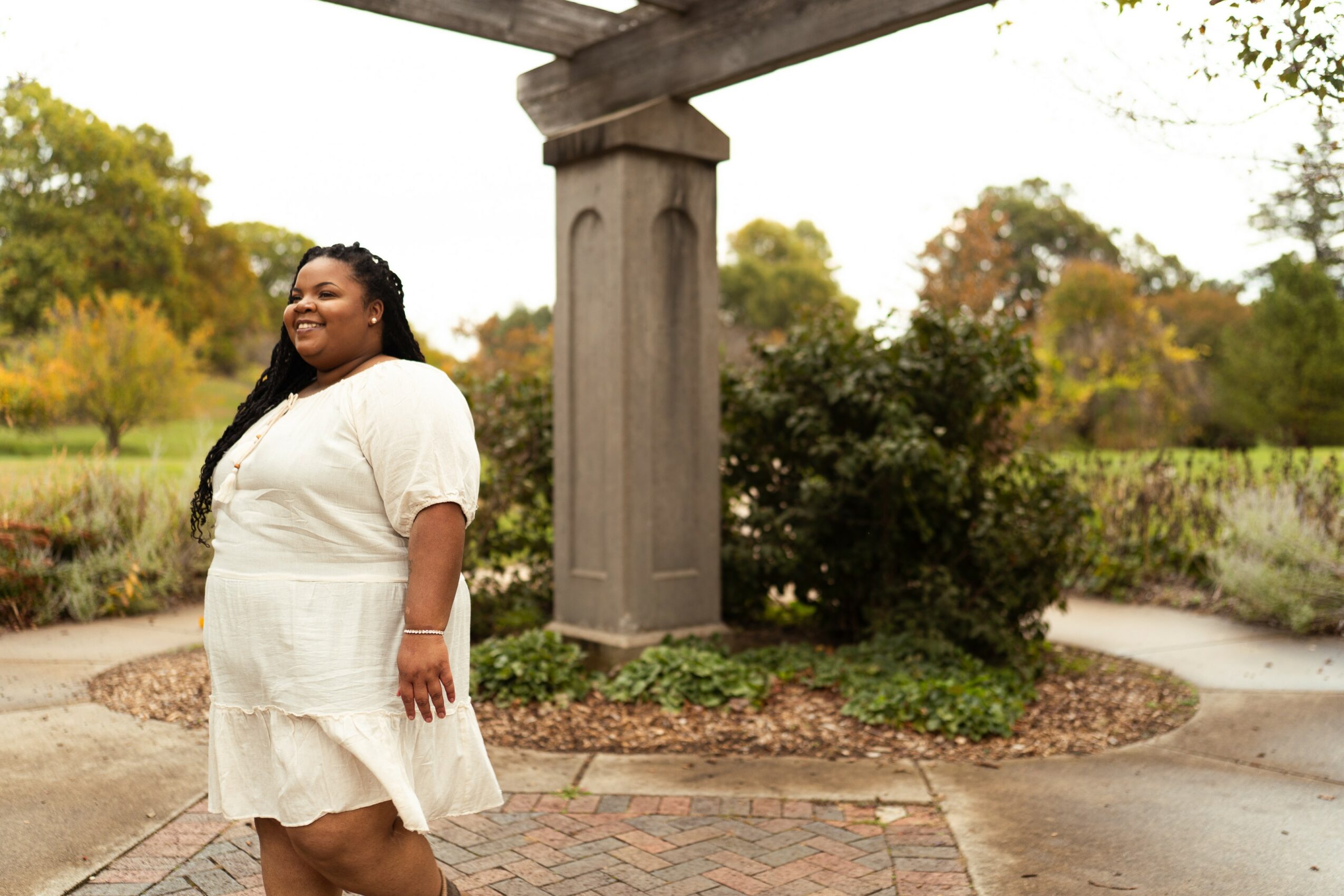 Smiling woman in white dress standing outdoors near a stone structure and greenery.