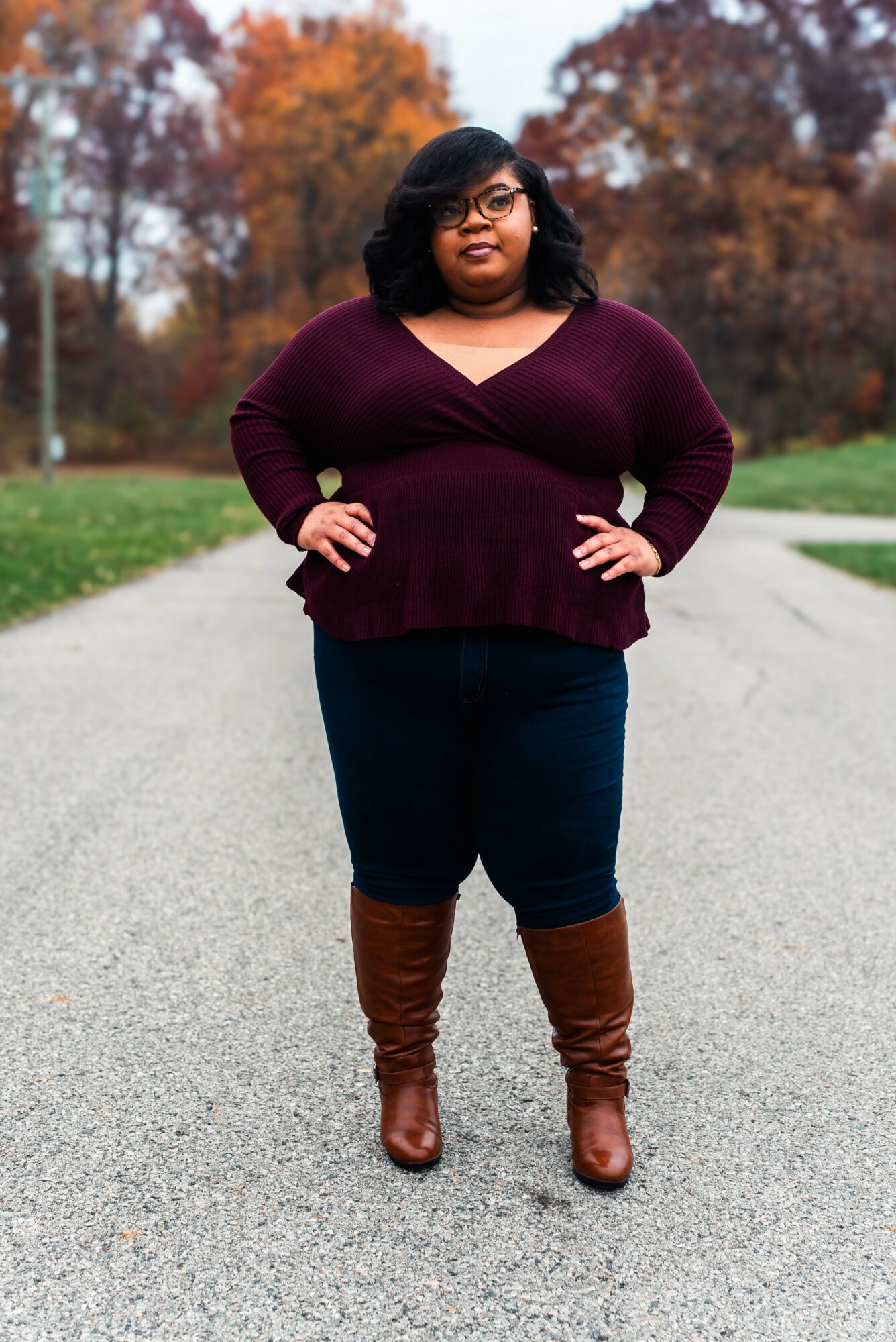 Woman standing outdoors on a paved path with autumn trees in the background, hands on hips, smiling.