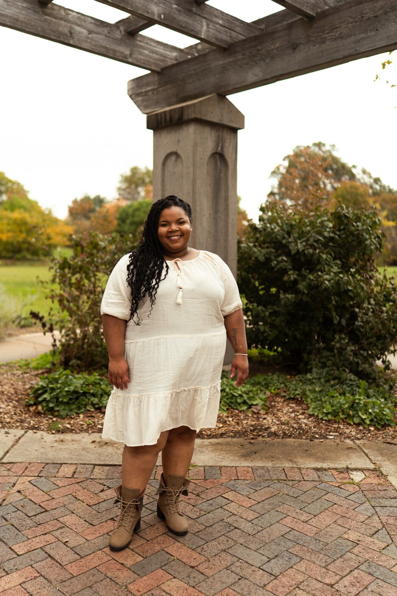 Woman standing outdoors under a wooden structure, smiling, wearing a white dress and boots, with trees and greenery in background.