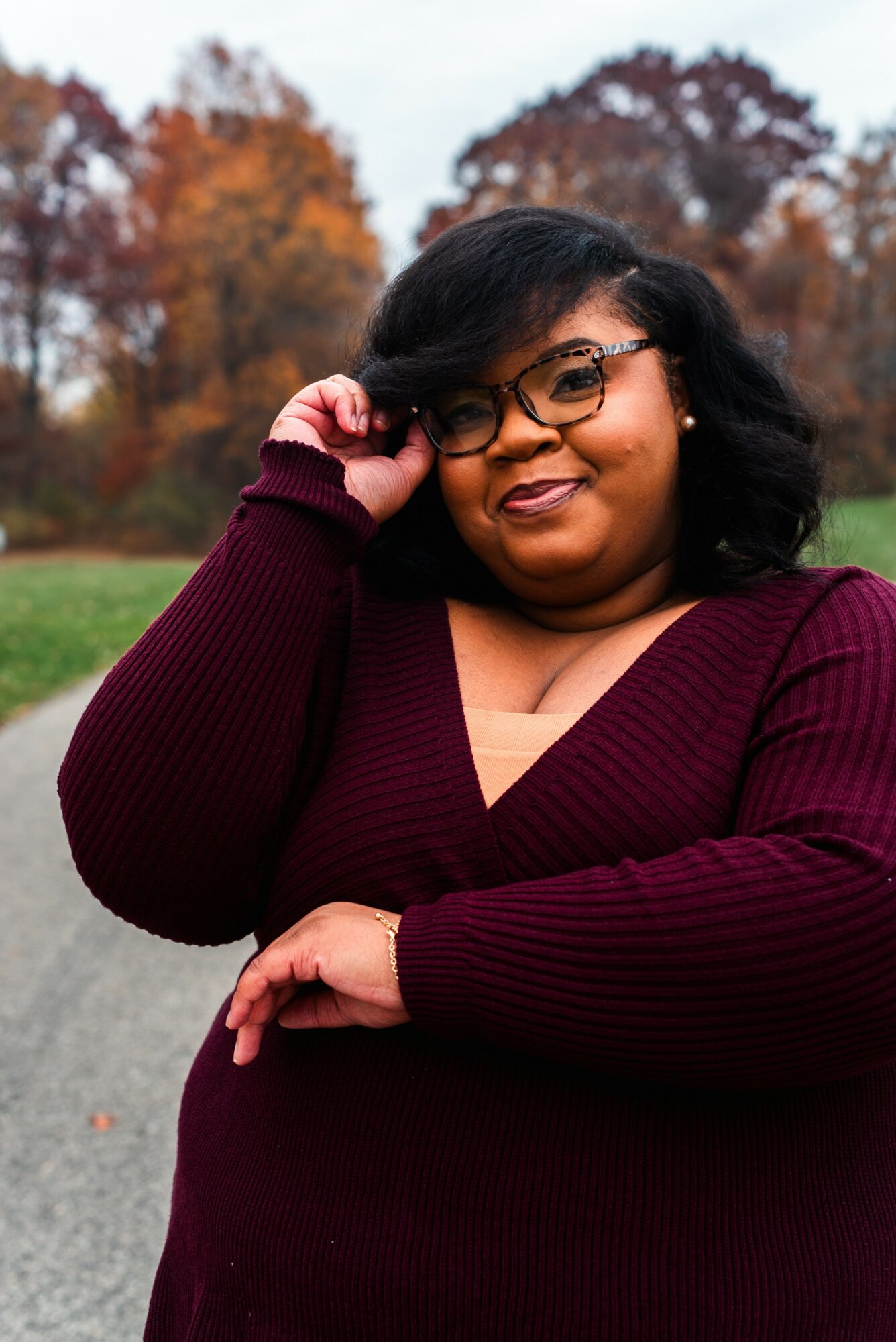 Woman with glasses and dark hair smiling outdoors in autumn, wearing a maroon sweater, trees with fall foliage in background.