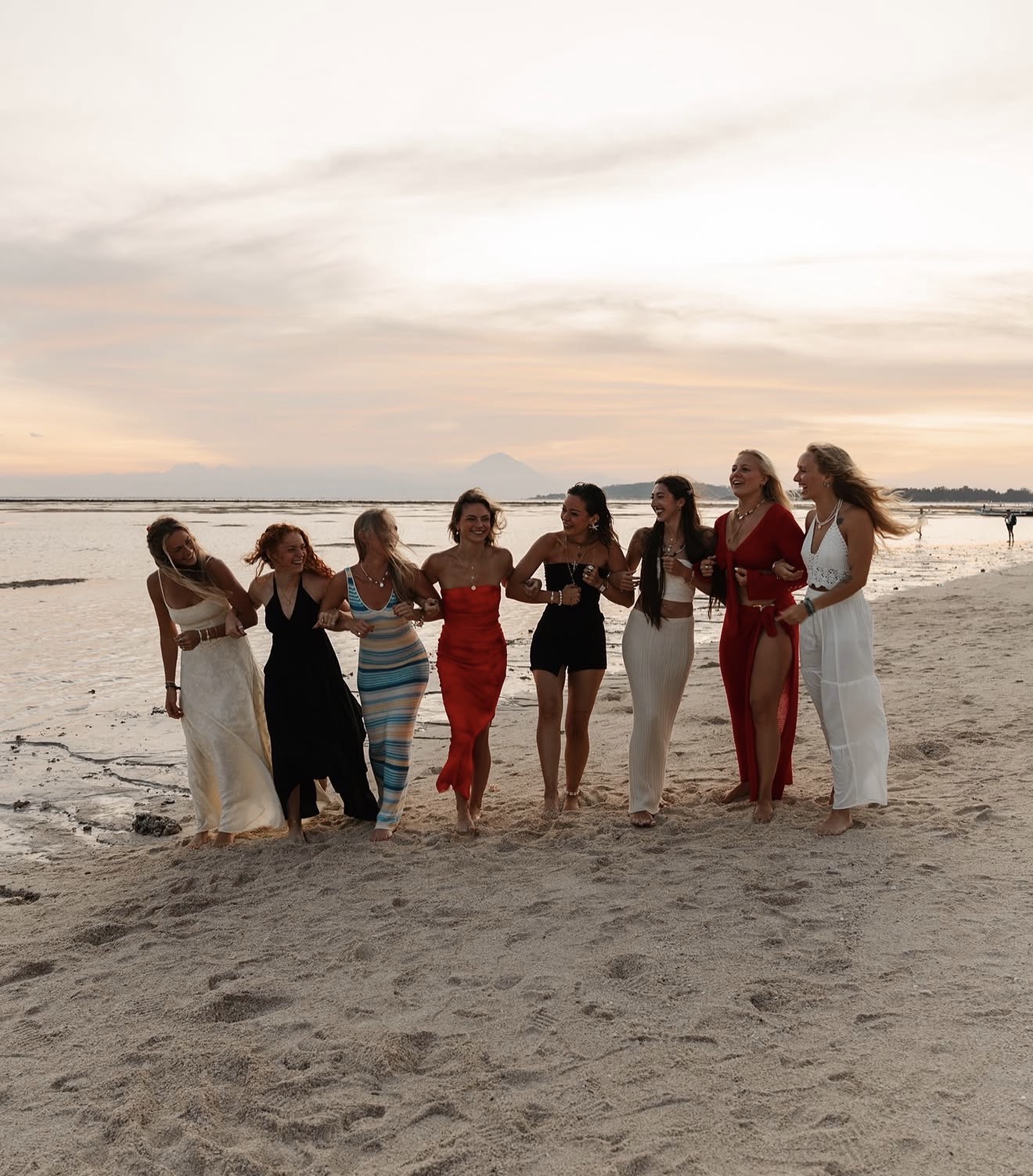 Group of nine women standing on a beach at sunset, wearing colorful dresses, smiling and talking.