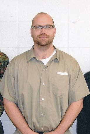Man with glasses and a beard wearing a beige shirt, standing against a white tiled wall.