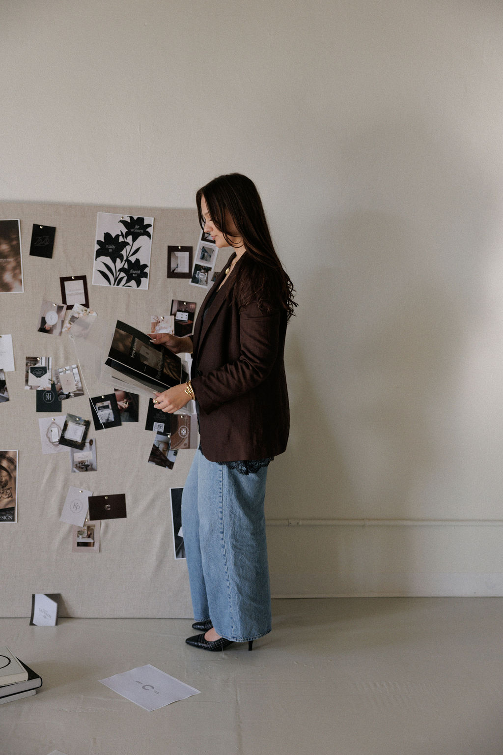 Woman with long hair, wearing a dark blazer and light jeans, stands reading a book in a room with a photo display on the wall.