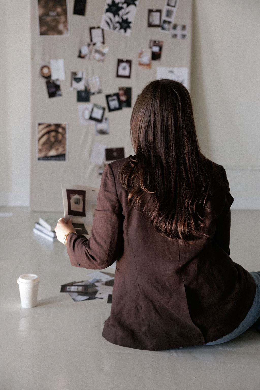 Person with long dark hair sitting on the floor, facing a wall with photos and pictures, holding a cup.