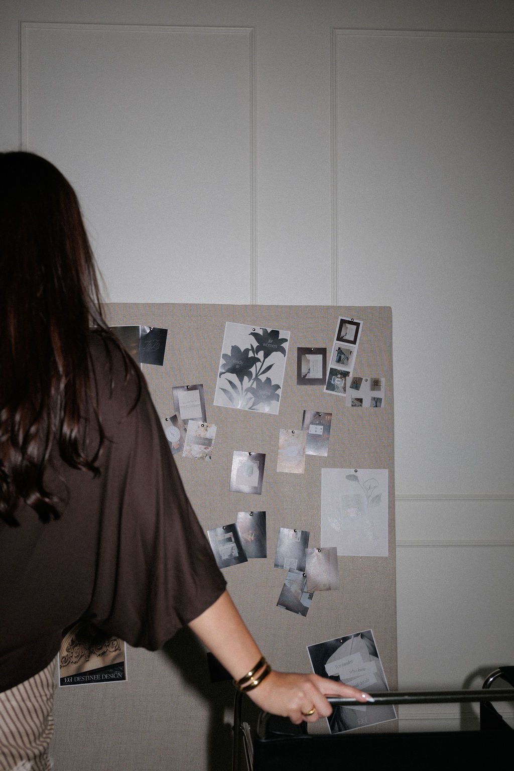 Person with long dark hair pointing at a bulletin board with photos and notes, in a room with white walls.