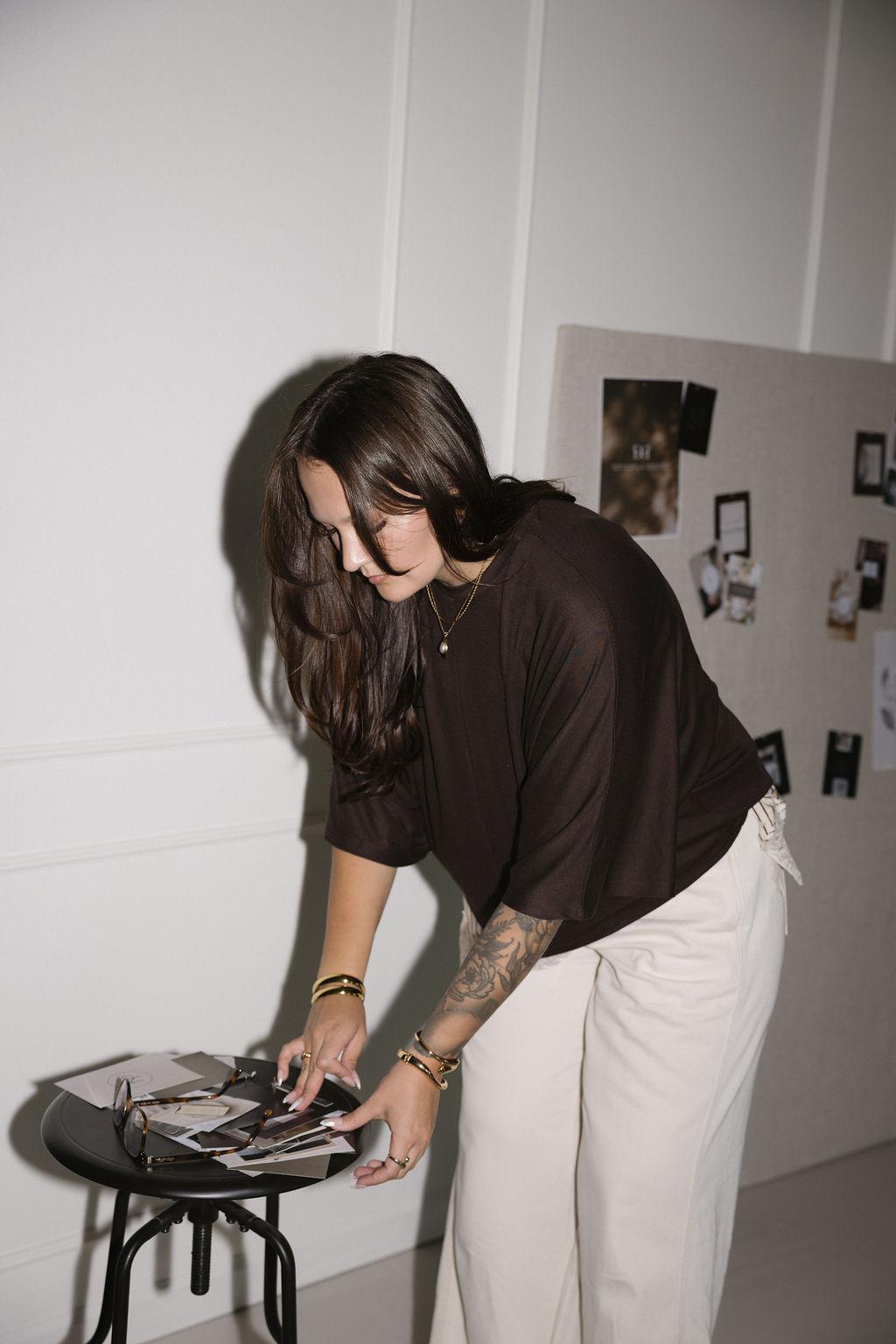 Woman with dark hair and tattoos, wearing a black top and white pants, arranging photos on a small table.