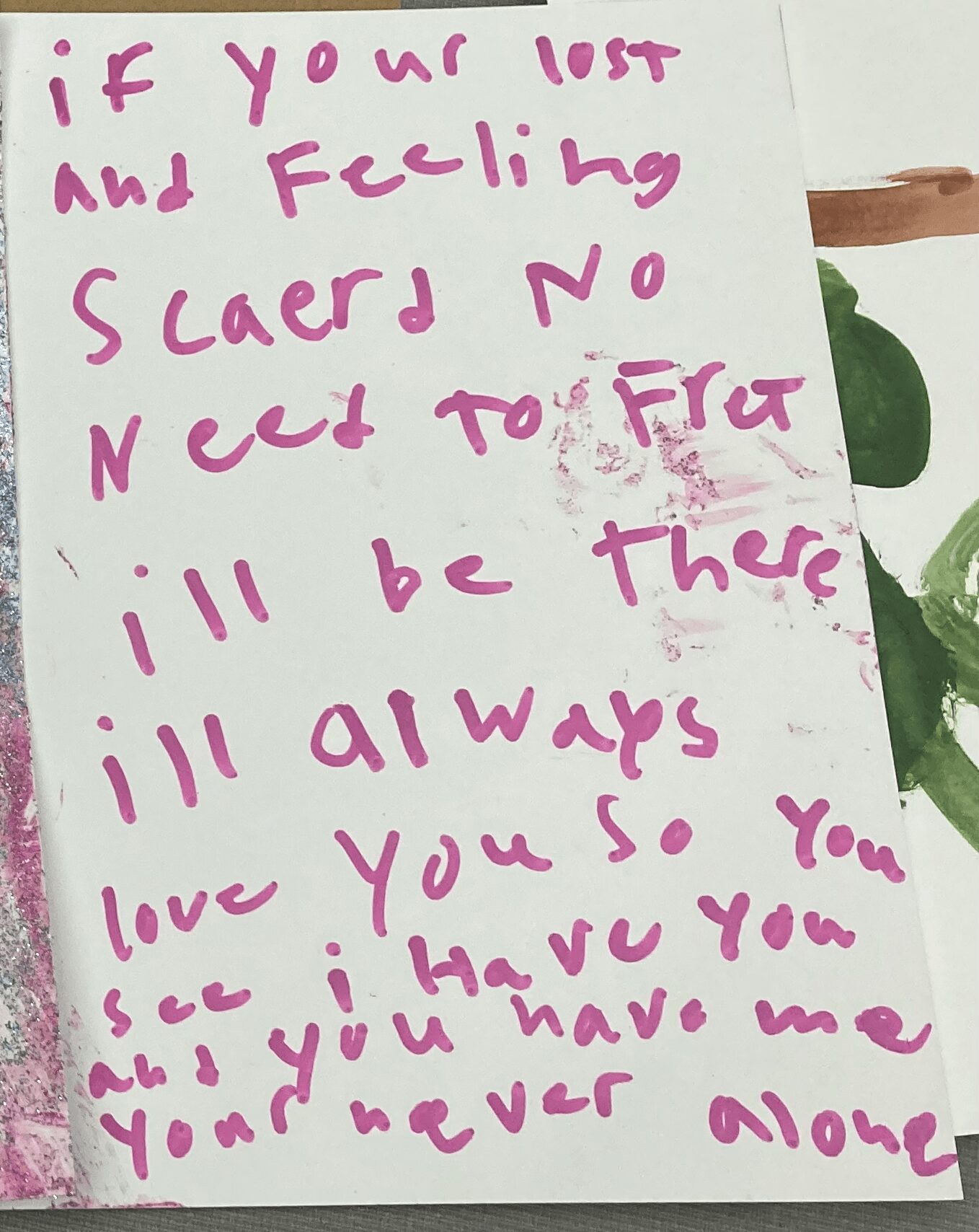 Handwritten note with pink ink on white paper, expressing love and support, with some smudges and a plant partially visible.