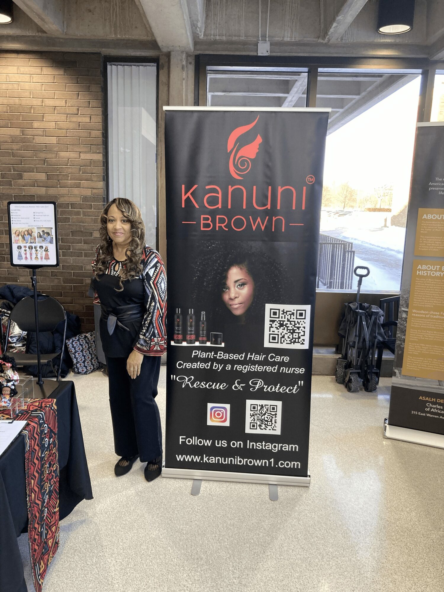 Woman standing next to a Kanuni Brown banner advertising hair care products, QR code, and social media info, in an indoor space.
