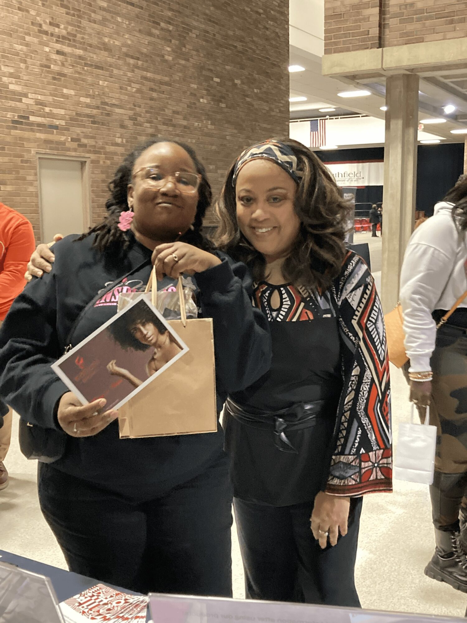 Two women standing indoors, smiling, one holding a photo and a paper bag, with a brick wall and other people in background.