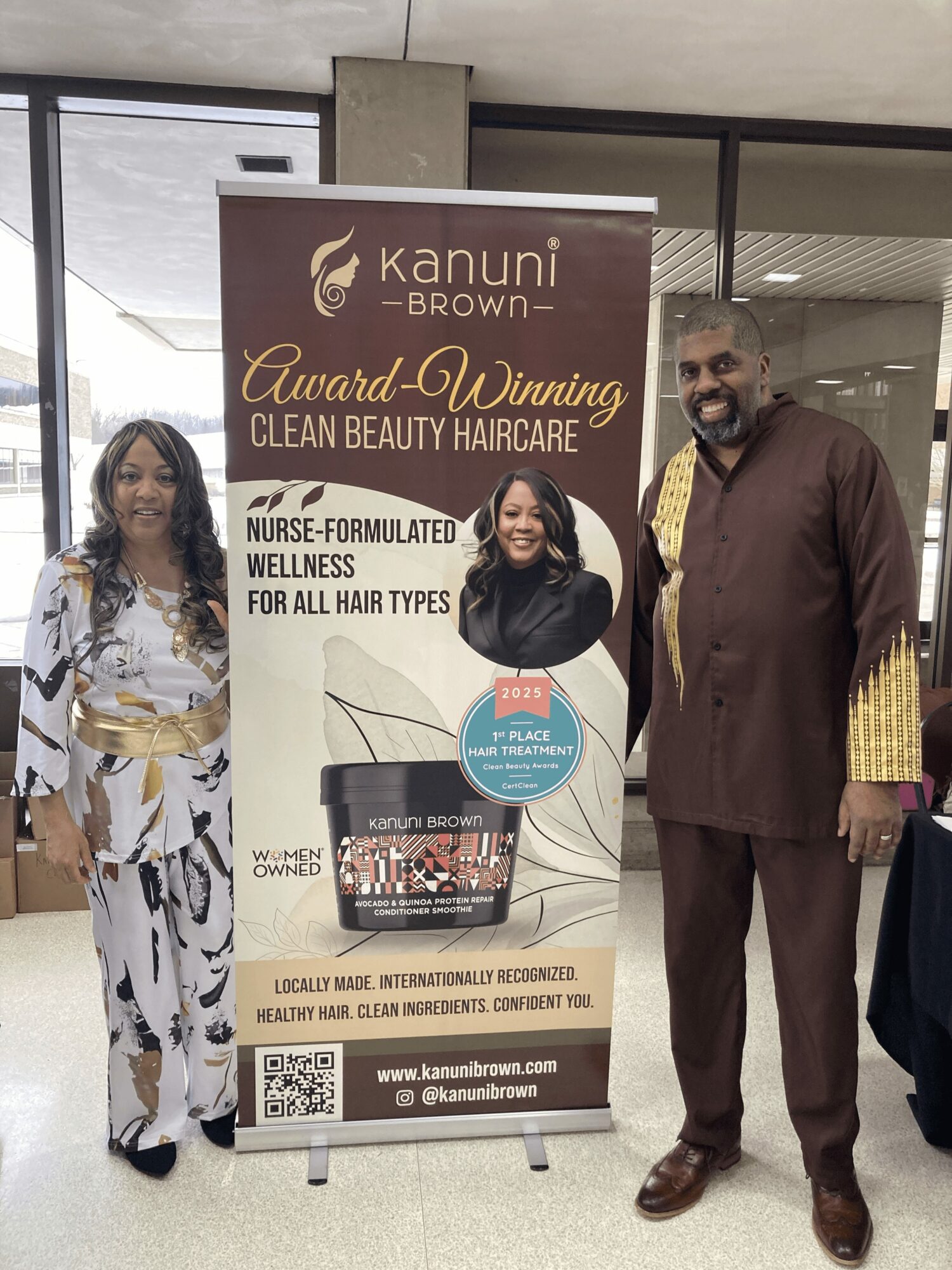 Three people standing next to a Kanuni Brown banner outside a building, smiling, with a woman in a white and yellow dress, a woman in black, and a man in brown attire.