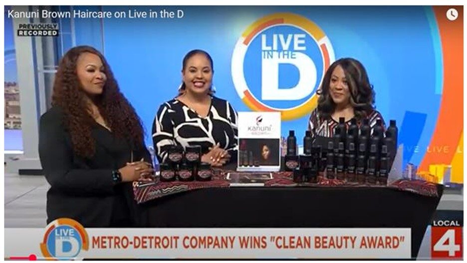 Three women stand behind a table with hair products, smiling, in a TV studio with a blue background and 'Live in the D' logo.