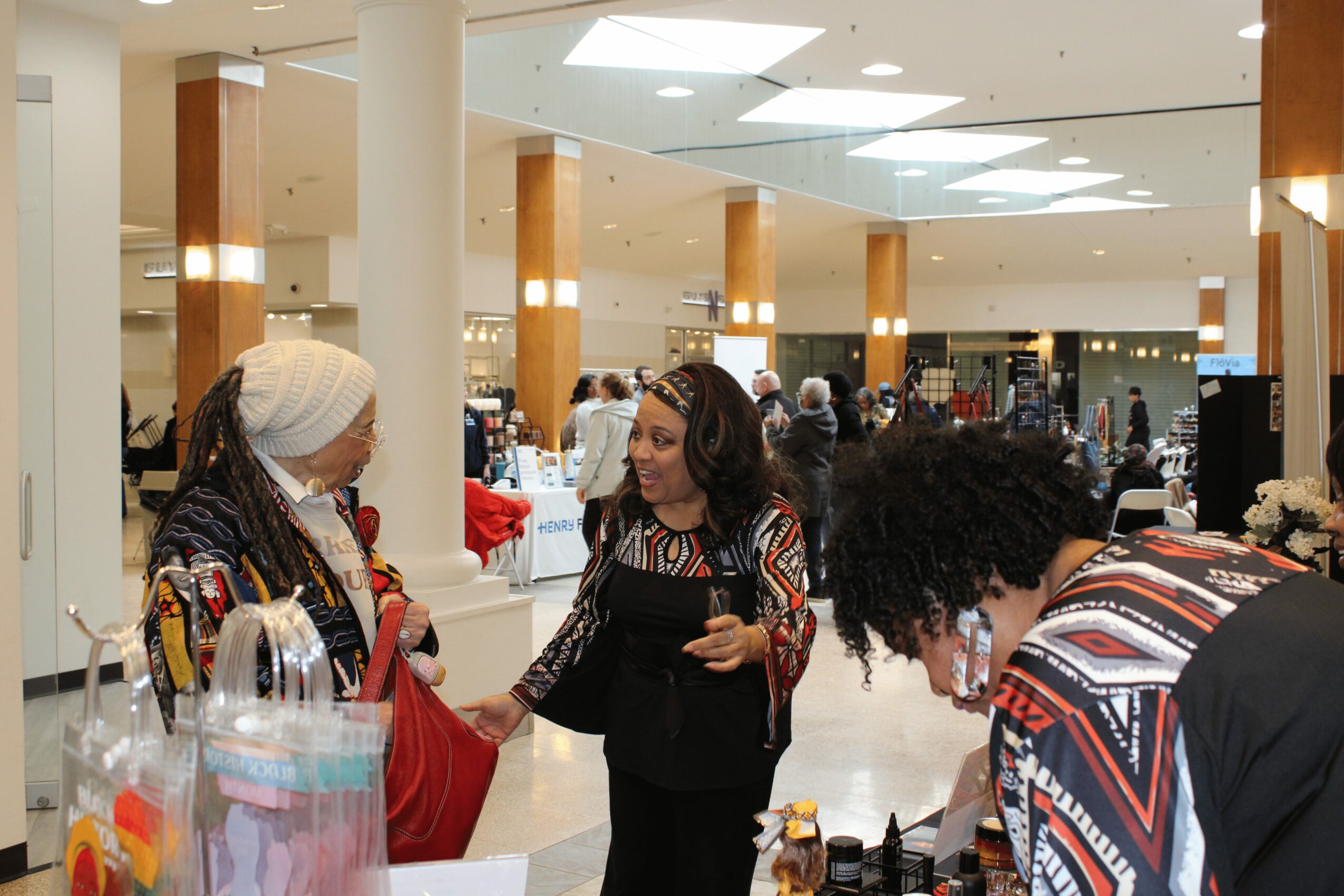 Three women engaged in conversation inside a shopping mall, with bright lighting and store displays in the background.