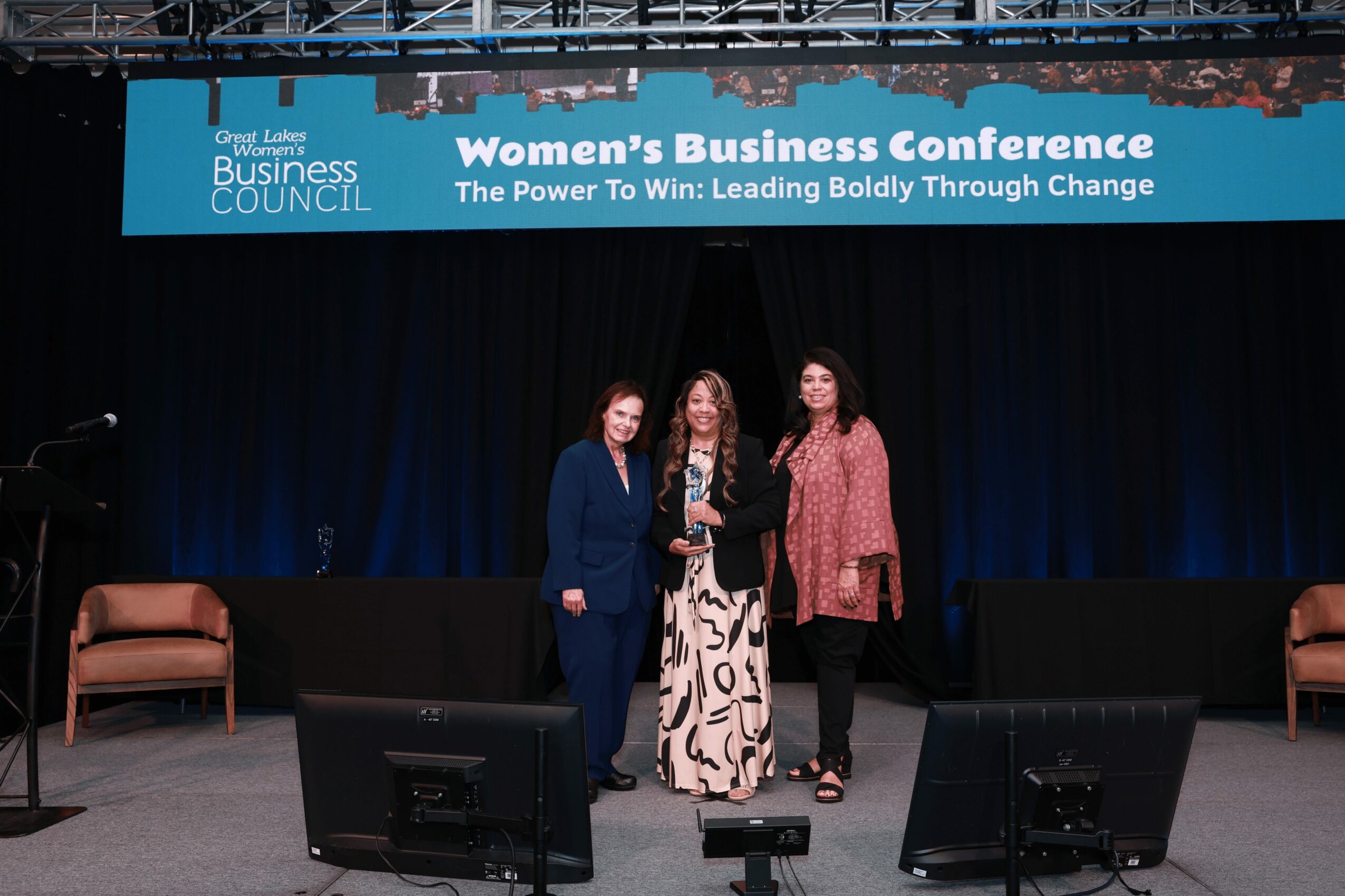 Three women standing on stage at a conference, with a banner above reading 'Women's Business Conference'.