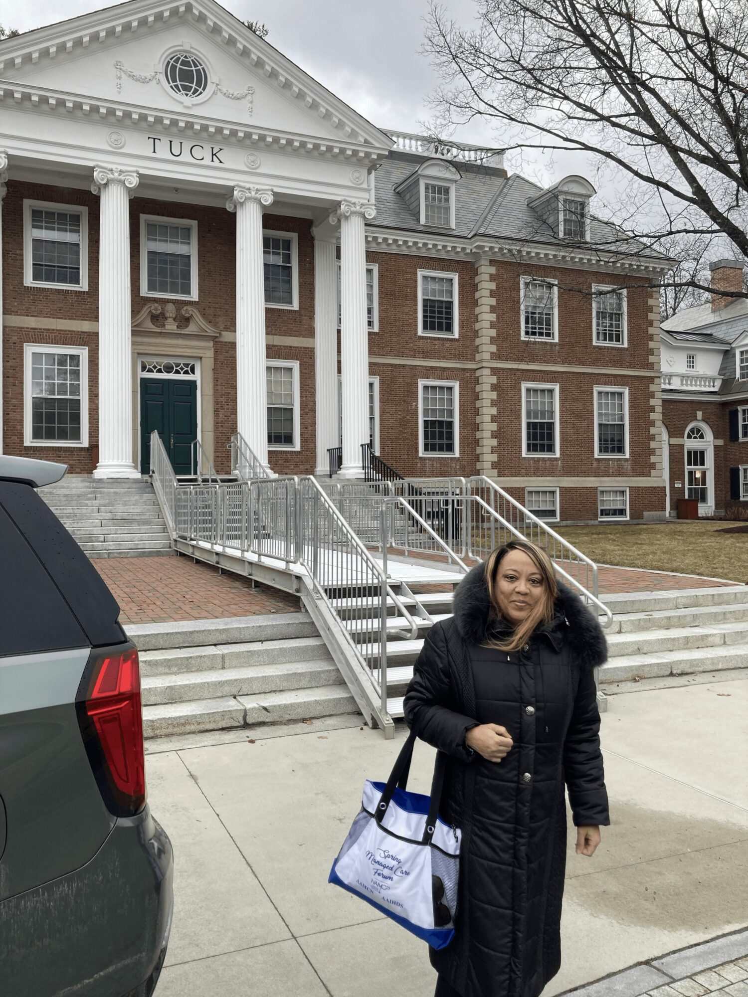 Woman standing outside a large brick building with white columns, holding a bag, smiling.