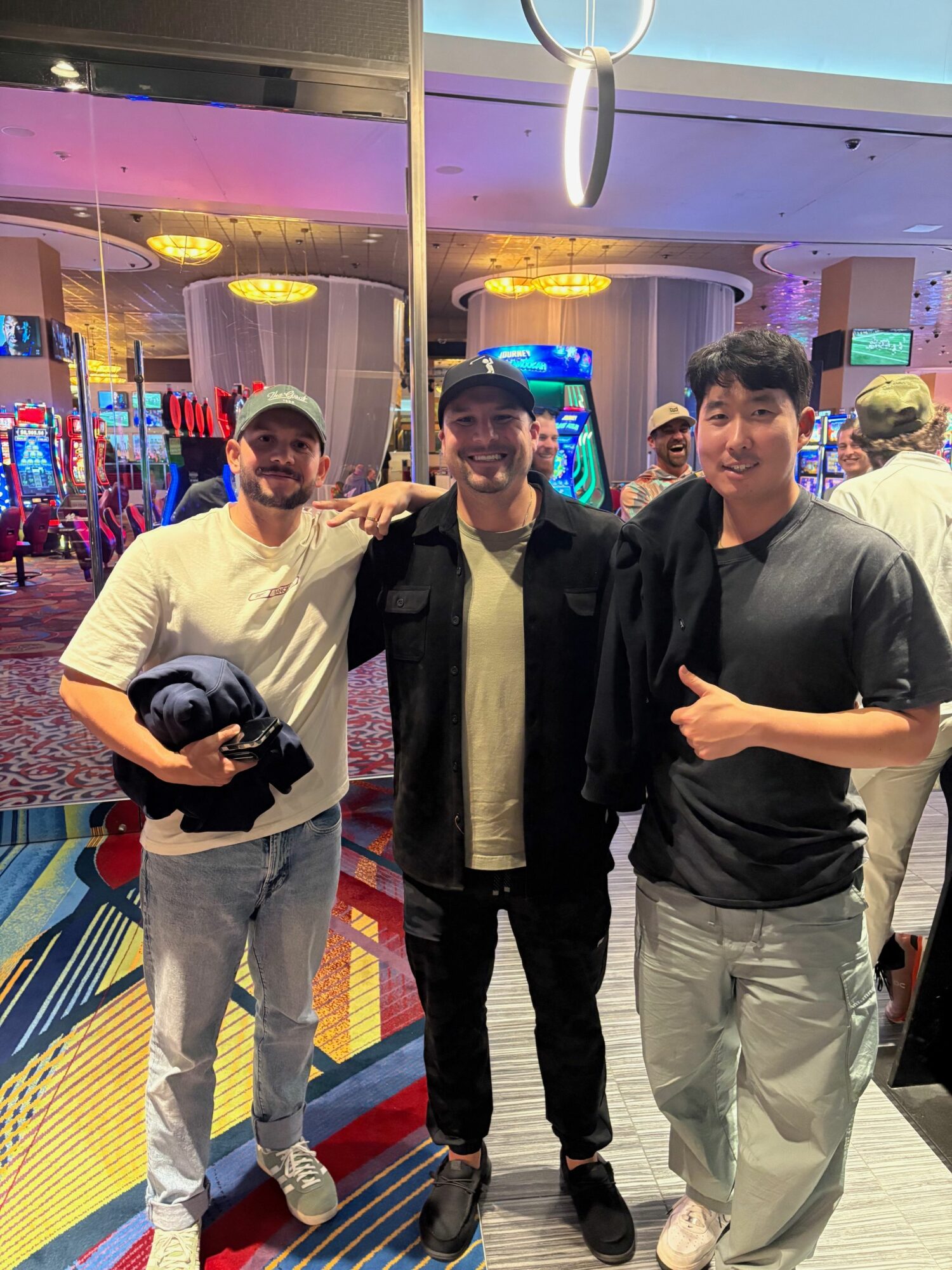 Three young men standing in an arcade, one holding a camera, with colorful gaming machines in background.