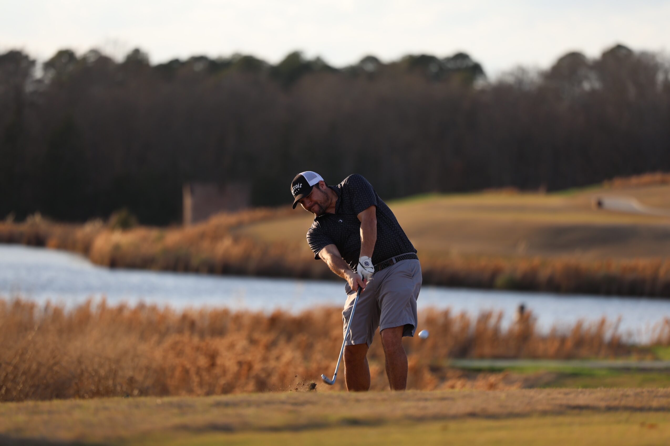 Person playing golf on a course near water, wearing a cap, dark shirt, and shorts, swinging a golf club.