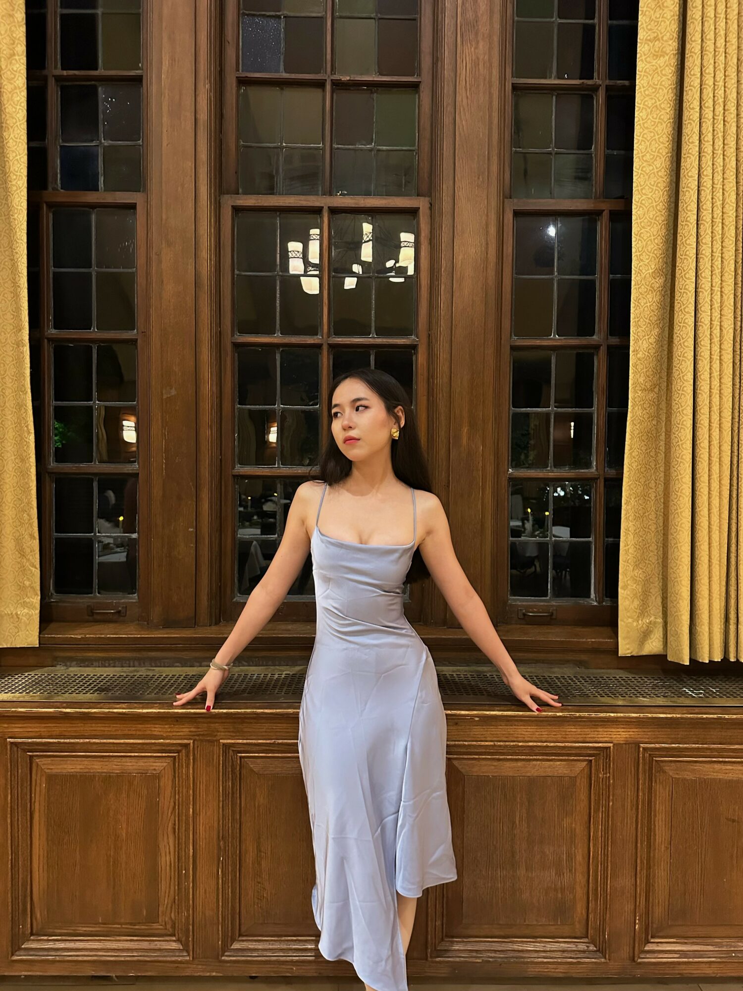 Woman in a light-colored dress standing with arms outstretched in front of large wooden window with yellow curtains.