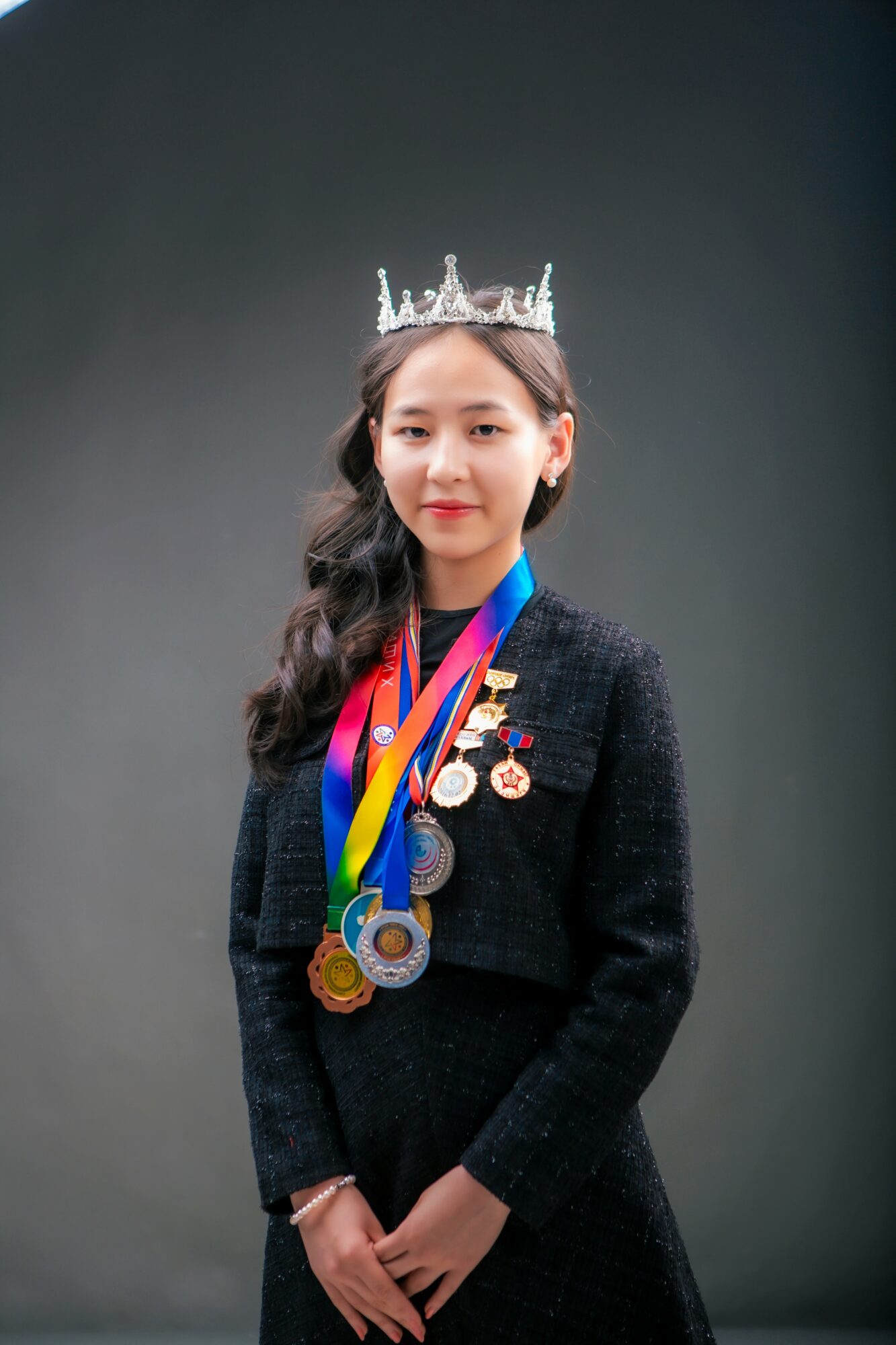 Young woman wearing a crown and medals, dressed in a dark outfit, standing against a plain background.