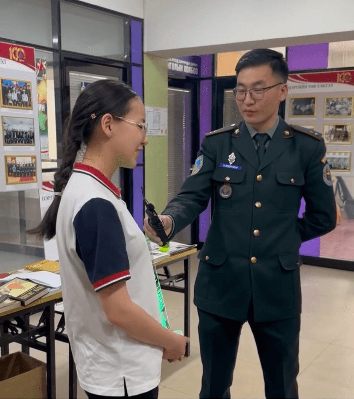 A young girl in a school uniform receives a medal from a man in a military uniform indoors, with display boards in the background.